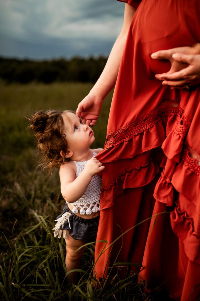 Maternity Photographer, baby daughter looks at expecting pregnant mother in a grass field