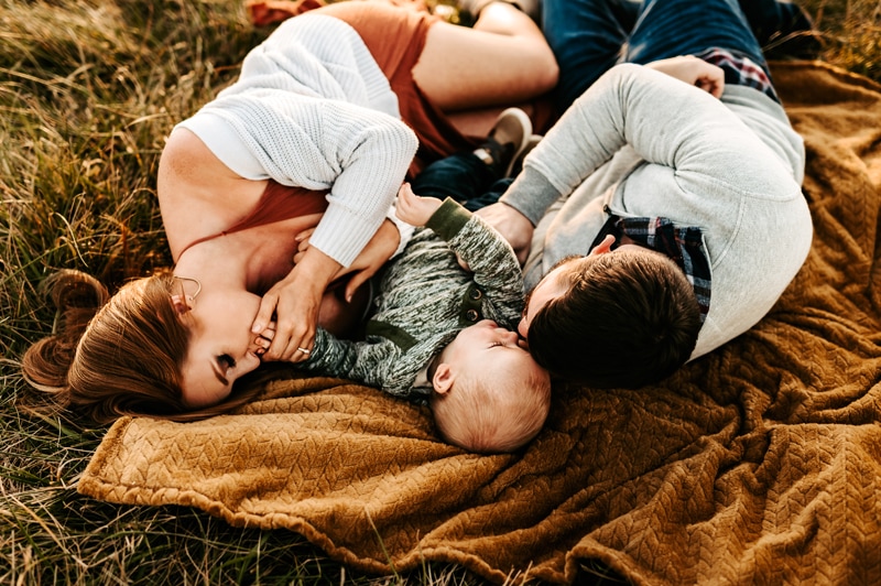 Family Photographer, mom and dad lay on blanket outside admiring baby